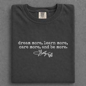 Puede incluir: Una camiseta gris carbón con cuello redondo y una etiqueta de Comfort Colors en el cuello. El frente presenta texto blanco que dice, dream more, learn more, care more, and be more, acompañado de una palabra en cursiva y dos mariposas.