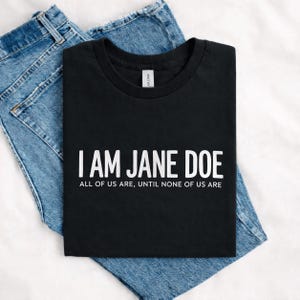 May include: A black short-sleeved t-shirt featuring the white text I AM JANE DOE followed by the text ALL OF US ARE, UNTIL NONE OF US ARE. The shirt is folded and placed on top of a pair of blue denim jeans with visible stitching and metal rivets.