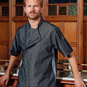 May include: A dark gray chef's jacket with short sleeves. The jacket has a unique asymmetrical design with a stand-up collar and a pocket on the left sleeve. The jacket has a gray panel on the side. The chef's jacket is made of a denim-like material.