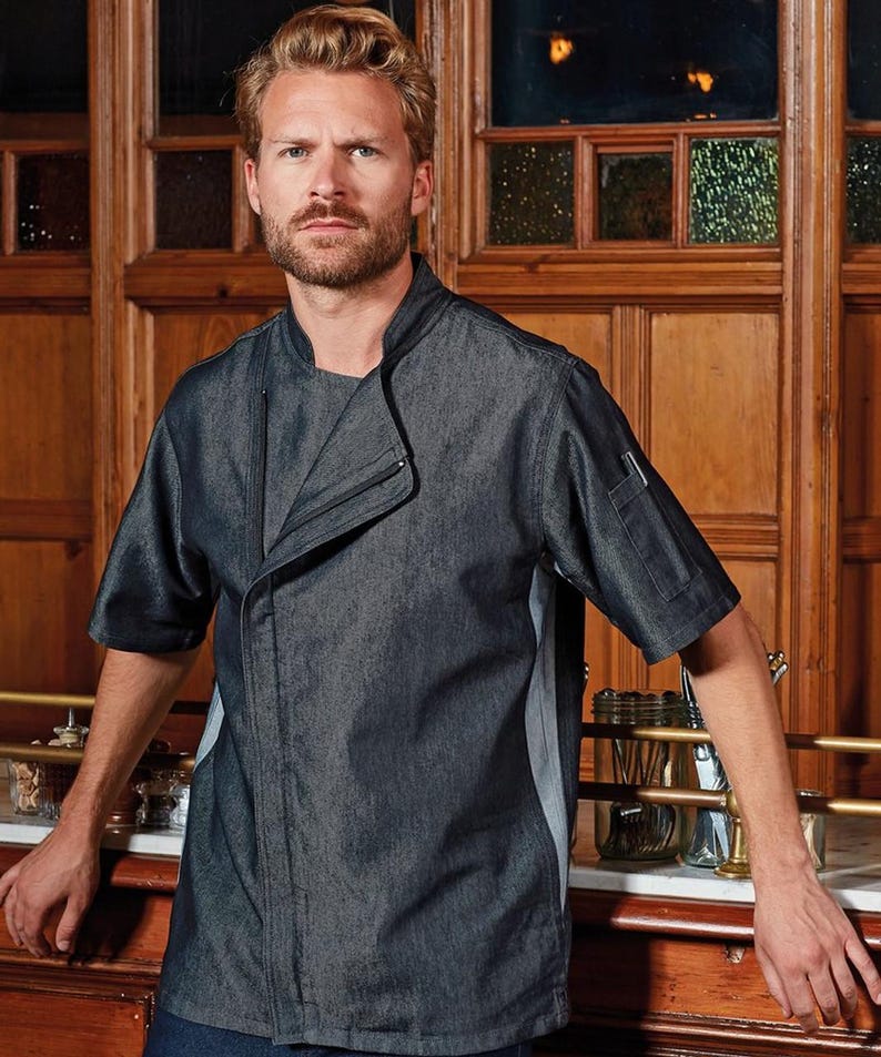 May include: A dark gray chef's jacket with short sleeves. The jacket has a unique asymmetrical design with a stand-up collar and a pocket on the left sleeve. The jacket has a gray panel on the side. The chef's jacket is made of a denim-like material.