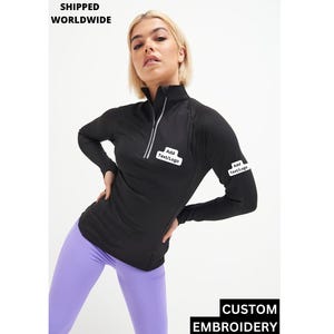 Puede incluir: Camiseta negra de manga larga con cuello alto y cremallera. La camiseta tiene parches blancos en la manga y el pecho que dicen "Añadir texto/logotipo". La modelo lleva la camiseta con leggings morados.