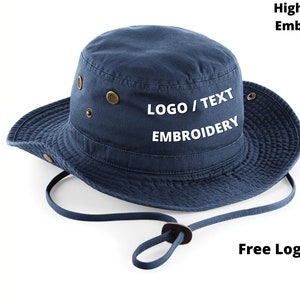 Puede incluir: Un sombrero de pescador azul marino con ala ancha y correa para la barbilla. El sombrero presenta ojales de color lat&oacute;n y el texto "LOGO / TEXT EMBROIDERY" en blanco. La imagen tambi&eacute;n incluye el texto "High Quality Embroidery" y "Free Logo Setup".