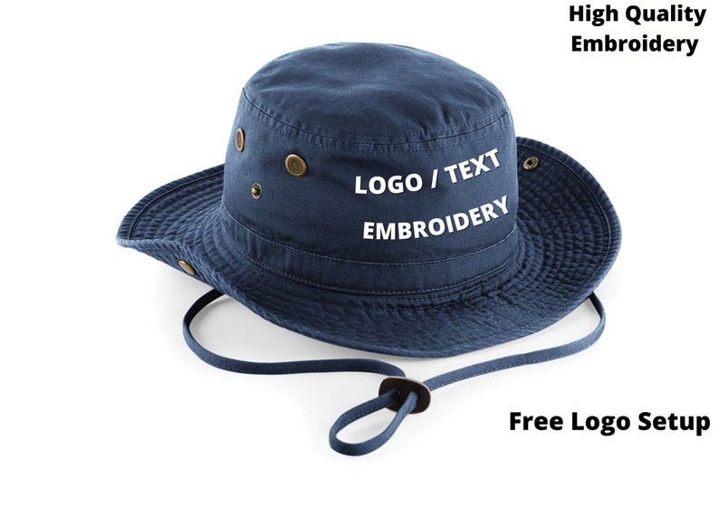 Puede incluir: Un sombrero de pescador azul marino con ala ancha y correa para la barbilla. El sombrero presenta ojales de color lat&oacute;n y el texto "LOGO / TEXT EMBROIDERY" en blanco. La imagen tambi&eacute;n incluye el texto "High Quality Embroidery" y "Free Logo Setup".