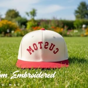 Könnte beinhalten: Cremefarbene Retro-Kappe mit rotem Schirm und rotem, gesticktem Schriftzug. Die Kappe liegt auf grünem Gras, mit einem verschwommenen Hintergrund aus Blumen und Bäumen. Der Text "Custom Embroidered Retro Hat" steht unten.