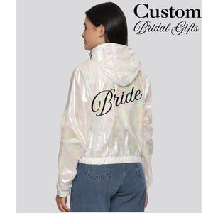May include: A white iridescent hooded jacket with the word "Bride" in black script on the back. The jacket is cropped and paired with blue jeans. Text at the top reads "Custom Bridal Gifts".