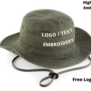 Puede incluir: Sombrero de pescador verde oliva con ala ancha y correa de barbilla ajustable. Cuenta con ojales met&aacute;licos y el texto "LOGO / TEXT EMBROIDERY" en blanco. El sombrero est&aacute; dise&ntilde;ado para bordado personalizado.