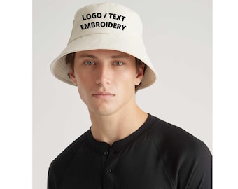 Puede incluir: Un sombrero de pescador color crema con el texto "LOGO / TEXT EMBROIDERY" en negro. El sombrero lo lleva una persona que lleva una camisa negra de manga larga. El sombrero es un accesorio informal.