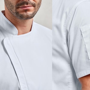 May include: Close-up of a white chef's coat. The coat features a mandarin collar, a diagonal closure, and a short sleeve with a small pocket. The fabric appears to be a crisp, durable material.