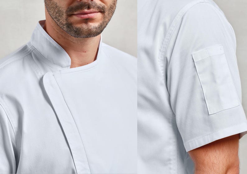 May include: Close-up of a white chef's coat. The coat features a mandarin collar, a diagonal closure, and a short sleeve with a small pocket. The fabric appears to be a crisp, durable material.