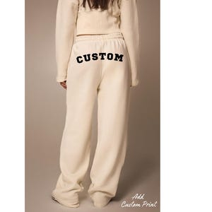 May include: Cream-colored sweatpants with the word "CUSTOM" printed in black across the back. The pants are part of a matching set, and the text "Add Custom Print" is visible in the lower right corner.