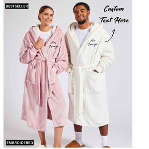 May include: Matching pink and white hooded bathrobes. The pink robe is worn by a person on the left, and the white robe is worn by a person on the right. Both robes have pockets, a belt, and the embroidered name "Mrs George" and "Mr George".