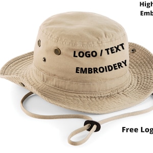 Puede incluir: Sombrero de pescador beige con ala ancha y correa de barbilla ajustable. El sombrero presenta ojales met&aacute;licos y el texto "LOGO / TEXT EMBROIDERY". La imagen tambi&eacute;n incluye el texto "High Quality Embroidery" y "Free Logo Setup".