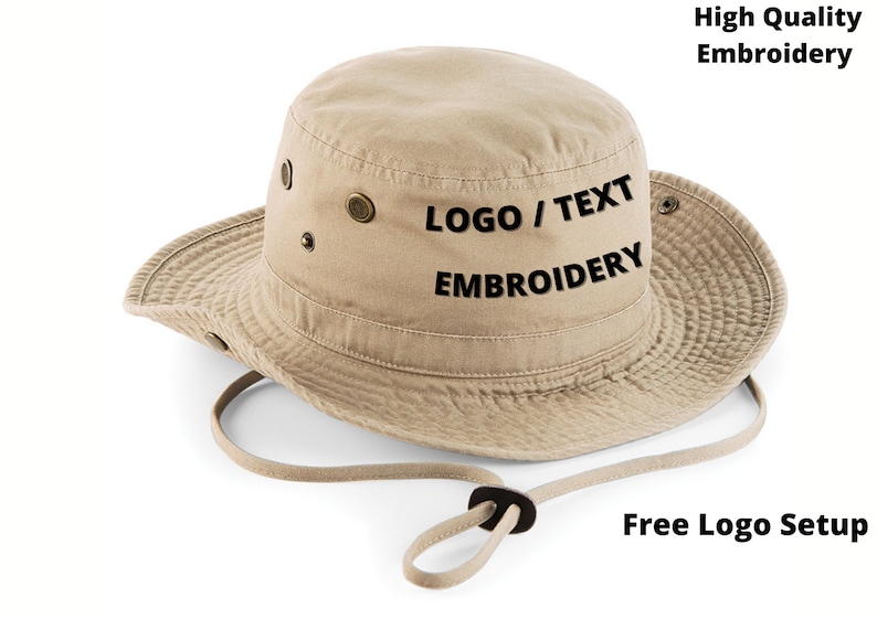 Puede incluir: Sombrero de pescador beige con ala ancha y correa de barbilla ajustable. El sombrero presenta ojales met&aacute;licos y el texto "LOGO / TEXT EMBROIDERY". La imagen tambi&eacute;n incluye el texto "High Quality Embroidery" y "Free Logo Setup".