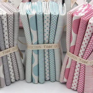 May include: Three bundles of fabric swatches in different colors and patterns. The bundles are tied with a brown ribbon and a label that reads "Collection Tilda Oui". The fabrics are in shades of gray, blue, and pink.