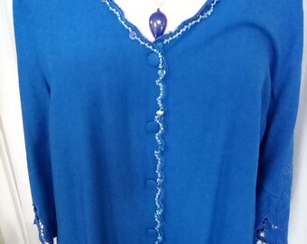 Puede incluir: Un cárdigan azul vibrante con cuello en V, adornado con delicados ribetes de cuentas y cierre de botones. Las mangas presentan un diseño de encaje floral en los puños. También es visible un collar plateado con un colgante azul oscuro.