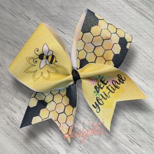 May include: A yellow and black cheer bow with a bee and honeycomb design. The bow has the text "Bee You-tiful" on it.