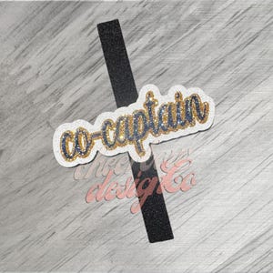 May include: A white sticker with the words "co-captain" in blue and gold glitter, with a black glitter stripe. The sticker is on a light gray wooden surface. The text "chyc designs co" is also visible.