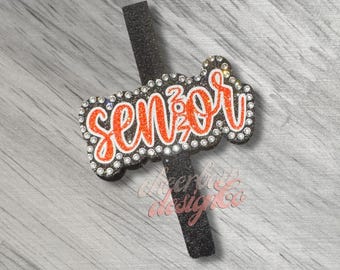 May include: A black glittery vertical strip attached to a horizontal black glittery plaque featuring the orange glittery script text "senior" outlined with small clear rhinestones. The entire piece is bordered by a row of clear rhinestones.