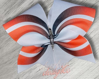 May include: A white cheer bow with orange and black stripes and a gradient design. The bow has a silver and rhinestone accent in the center.
