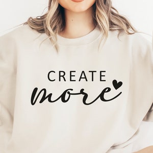 May include: A white sweatshirt with the text "CREATE more" in black script with a small black heart. The model is wearing the sweatshirt and a white beanie and is making the peace sign with both hands.