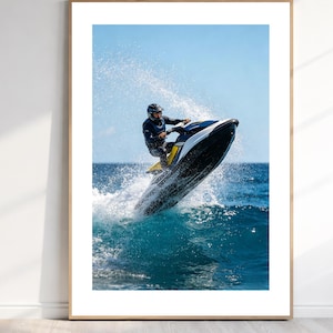 May include: A framed wall art print featuring a jet ski jumping over a blue ocean wave. The jet ski is black, white, and yellow, and the rider wears a dark wetsuit and helmet. The scene captures a high-speed water action moment with spray.