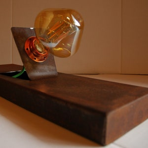 May include: A rustic metal lamp base with a single light bulb. The base is made of dark brown metal with a rectangular shape. The light bulb is a clear glass bulb with a filament visible inside. The lamp is on a white surface.