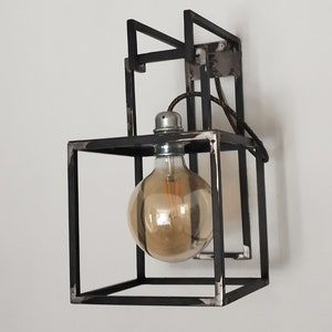 Może przedstawiać: Czarna metalowa lampa ścienna o geometrycznym wzorze. Lampa posiada jedną żarówkę z przezroczystym kloszem.