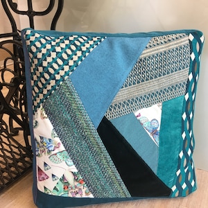 Könnte beinhalten: Ein blaugrünes Patchwork-Kissen mit geometrischem Muster. Das Kissen besteht aus verschiedenen Stoffen und Texturen, darunter Samt, Baumwolle und Leinen.