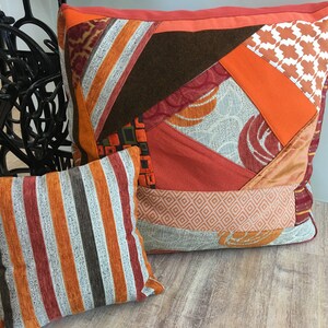 Könnte beinhalten: Zwei dekorative Zierkissen. Das größere Kissen hat ein Patchwork-Design mit orangefarbenen, braunen und roten Stoffen. Das kleinere Kissen hat vertikale Streifen in Orange, Braun und Weiß. Beide Kissen liegen auf einer Holzoberfläche.