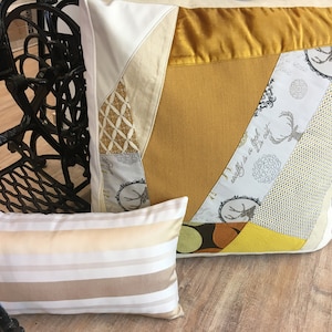 Könnte beinhalten: Zwei dekorative Kissen mit Patchwork-Muster. Das größere Kissen hat ein gelbes, weißes und graues Muster mit einem Hirschmotiv. Das kleinere Kissen hat ein weiß-beiges Streifenmuster.