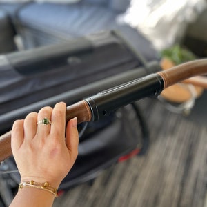 Pu&ograve; includere: Una mano che tiene la maniglia in pelle marrone di un passeggino nero con un anello d'argento al dito.