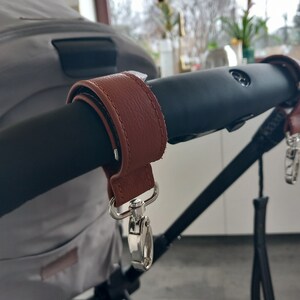 May include: A brown leather stroller handle wrap with a silver clip. The wrap is attached to the black stroller handle.