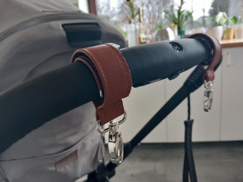 May include: A brown leather stroller handle wrap with a silver clip. The wrap is attached to the black stroller handle.