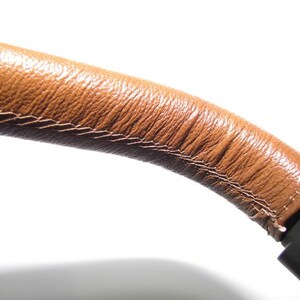 May include: Close-up of a brown leather strap with stitching detail. The strap is attached to a black object.