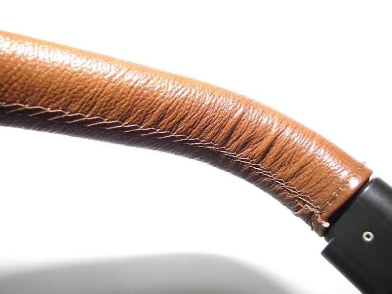 May include: Close-up of a brown leather strap with stitching detail. The strap is attached to a black object.