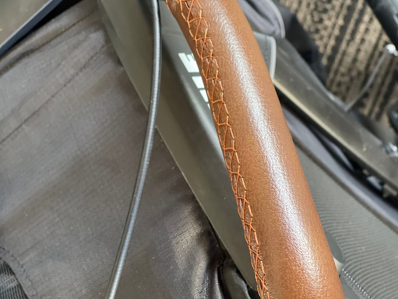 Pu&ograve; includere: Primo piano di un manubrio in pelle marrone con cuciture arancioni. Le cuciture sono un dettaglio decorativo sul manubrio.