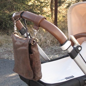 May include: A brown leather bag hanging from the handle of a stroller. The stroller has a white fabric seat and a brown leather handlebar.