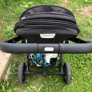 Op de afbeelding: Een zwarte kinderwagen met een zwart leren handvat en een zwarte kap. De kinderwagen heeft een zwarte mand eronder met een kleurrijk bloemenpatroon.