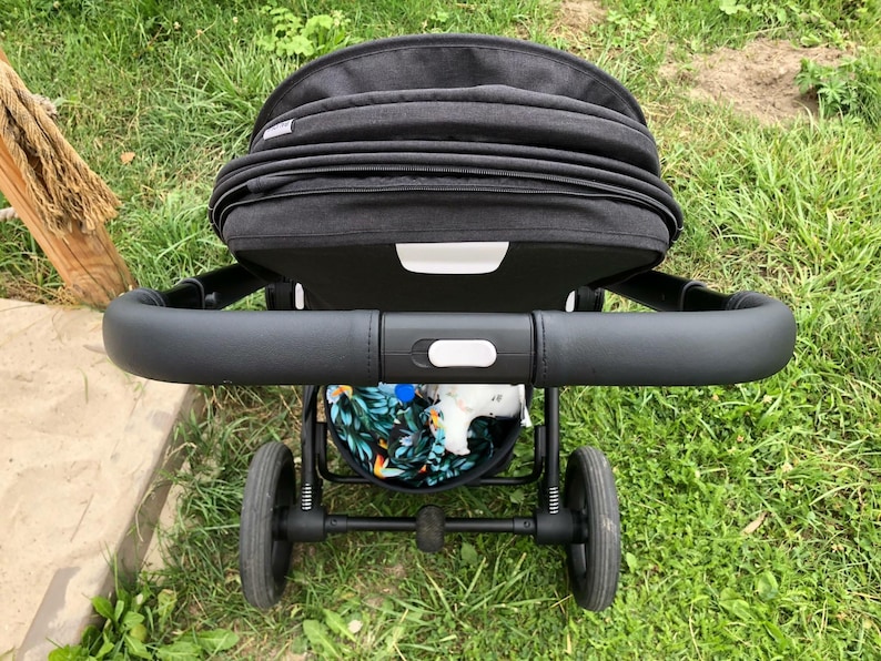 Op de afbeelding: Een zwarte kinderwagen met een zwart leren handvat en een zwarte kap. De kinderwagen heeft een zwarte mand eronder met een kleurrijk bloemenpatroon.