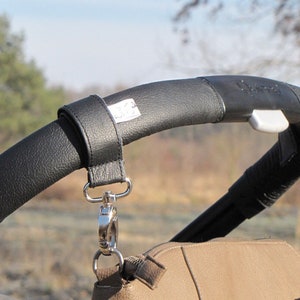 May include: Black leather stroller handlebar strap with silver clips. The strap is attached to a brown leather bag.