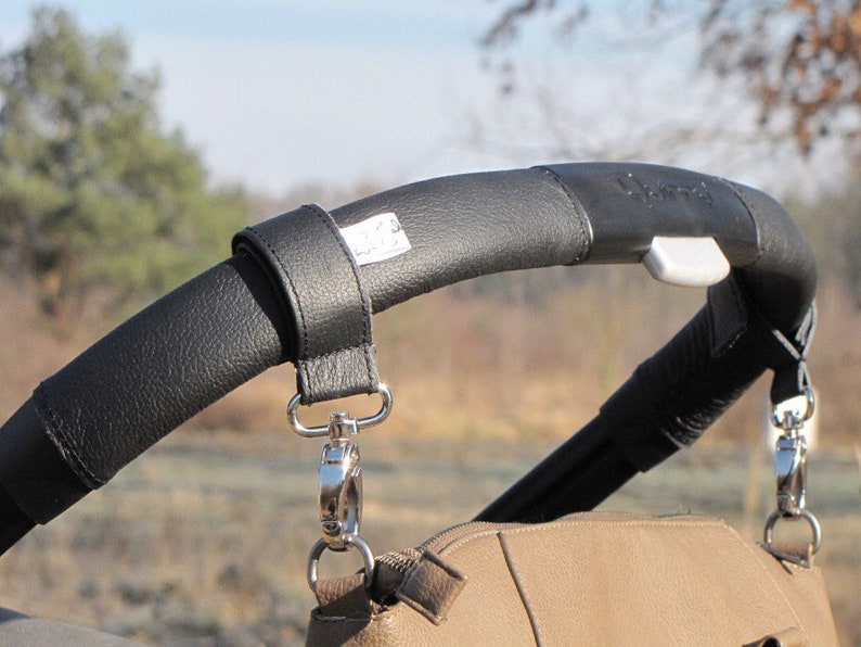 May include: Black leather stroller handlebar strap with silver clips. The strap is attached to a brown leather bag.