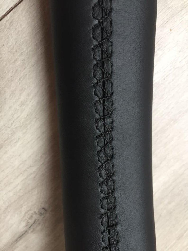 May include: Close-up of a black leather steering wheel cover with black stitching detail.