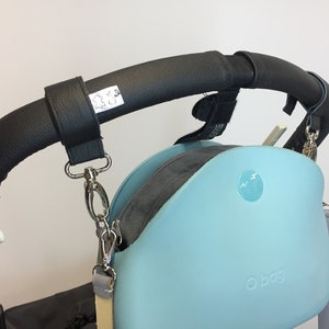 May include: A black leather stroller handlebar strap with a silver clip attached to a light blue O bag with a zipper and a silver chain.