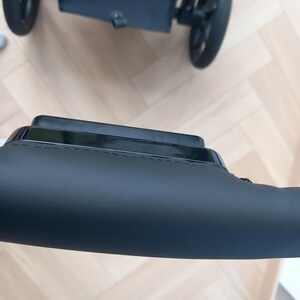 May include: A black stroller handlebar with a leather grip. The handlebar is made of metal and has a black finish. The leather grip is soft and comfortable to hold.