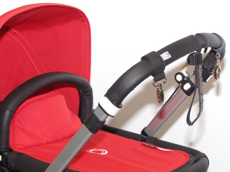 May include: A red stroller with a black leather handlebar and a black leather handle on the side. The stroller has a white strap with a silver buckle and a black strap with a silver buckle.