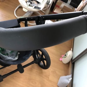 Op de afbeelding: Zwarte kinderwagen met een gevoerd stuur en een zwarte mand eronder. De kinderwagen staat op een houten vloer.
