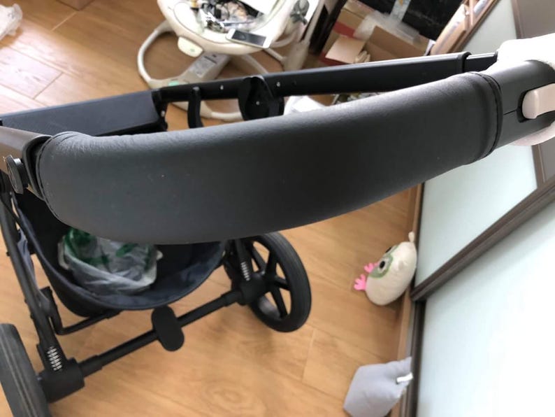 Op de afbeelding: Zwarte kinderwagen met een gevoerd stuur en een zwarte mand eronder. De kinderwagen staat op een houten vloer.