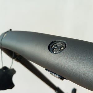 May include: Close-up of a grey leather handlebar with a black logo. The logo is a stylised letter "F" with a curved design.