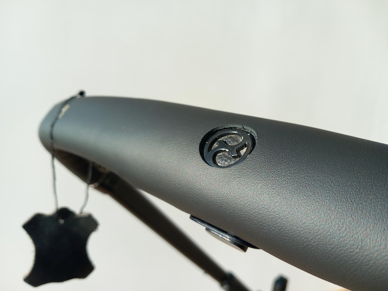 May include: Close-up of a grey leather handlebar with a black logo. The logo is a stylised letter "F" with a curved design.