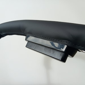 May include: Close-up of a black leather steering wheel cover with a black plastic control panel attached.